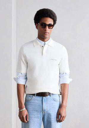 Homme portant des lunettes de soleil foncées, un polo blanc à manches courtes sur une chemise rayée bleue, un jean bleu clair et une ceinture noire, debout contre un mur uni.