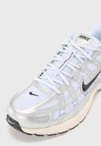 Běžecká obuv Nike s bílým síťovaným svrškem se stříbrnými akcenty, černým logem a texturovanou gumovou podrážkou. Šněrovací tkaničky jsou bílé s visačkou.