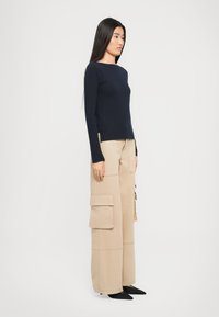 Femeie cu păr lung și negru, îmbrăcată cu un top lung de mătase de culoare bleumarin, pantaloni cargo bej cu picioare largi și pantofi negri cu vârful ascuțit, stând în profil.