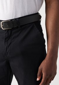 Curea neagră din piele cu cataramă argintie, purtată cu pantaloni negri. Textura materialului pare netedă; curea are găuri vizibile și ajustare pentru un fit personalizat.