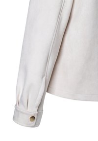 Ärmel einer hellbeigen Wildlederjacke mit einem durch einen einzelnen goldenen Druckknopf befestigten Bündchen und sichtbaren Nahtdetails.