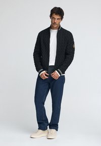 Svart kabelstickad kofta med vita randiga muddar och krage, kombinerad med blå jeans och ljusa sneakers. Modellen står mot en enfärgad bakgrund.