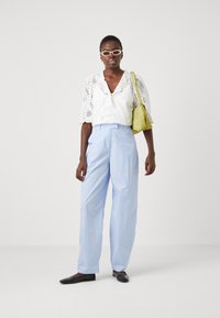 Witte blouse met oogjes en pofmouwen, lichtblauwe gestreepte hoge taillebroek, en een groene handtas. Zwarte platte schoenen maken de outfit compleet.
