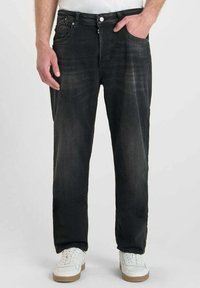 Jean en denim noir à coupe droite, avec des détails déchirés, un design à cinq poches et une fermeture à bouton, portés avec des baskets blanches.