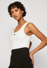 Valge ribadega tank tops laia rihmaga, millel on must kolmnurkne logoaktsent. Sobib kõrge vöökohaga mustade pükstega, millel on nupud.