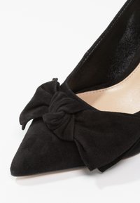 Chaussure noire en daim à bout pointu avec un grand nœud noué sur le devant et une doublure intérieure beige.