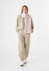 Een jonge vrouw in een beige trainingsbroek en sweatshirt, witte sneakers en een dikke roze sjaal, staand tegen een witte achtergrond.