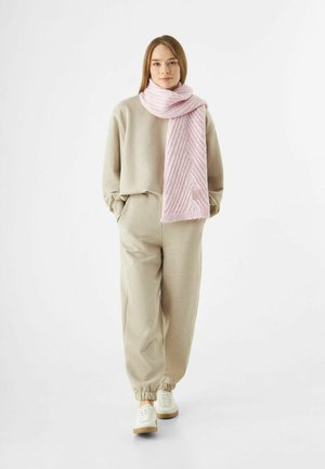 Junge Frau in beigen Sweatpants und Sweatshirt, weißen Sneakers und einem dicken rosa Schal, der vor einem weißen Hintergrund steht.