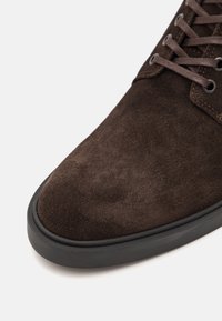 Cizmă din suede maro, în prim-plan, cu bot rotunjit, talpă din cauciuc negru și șireturi maro asortate. Textură moale, cu accente minime.