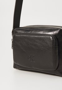 Čierna kožená crossbody taška s textúrovaným povrchom, obsahujúca zipsovú prednú vrecko a embosované logo. Súčasťou je nastaviteľný popruh.