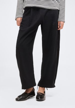 Osoba nosiaca voľné čierne nohavice, čierne baleríny na šnurovanie a dlhý rukáv, čierno-biele pruhované tričko, stojaca na jednofarebnom pozadí.