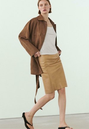 Kvinde, der går iført brun oversized jakke, hvid tanktop, beige knælængde nederdel og sorte sandaler mod en ensartet lys baggrund.