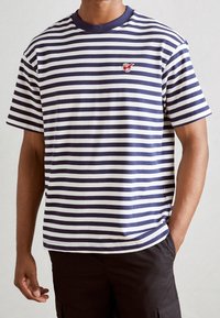 Stribet t-shirt med marineblå og hvide vandrette striber, ribbet marineblå krave og et lille rødt emblem på venstre bryst.