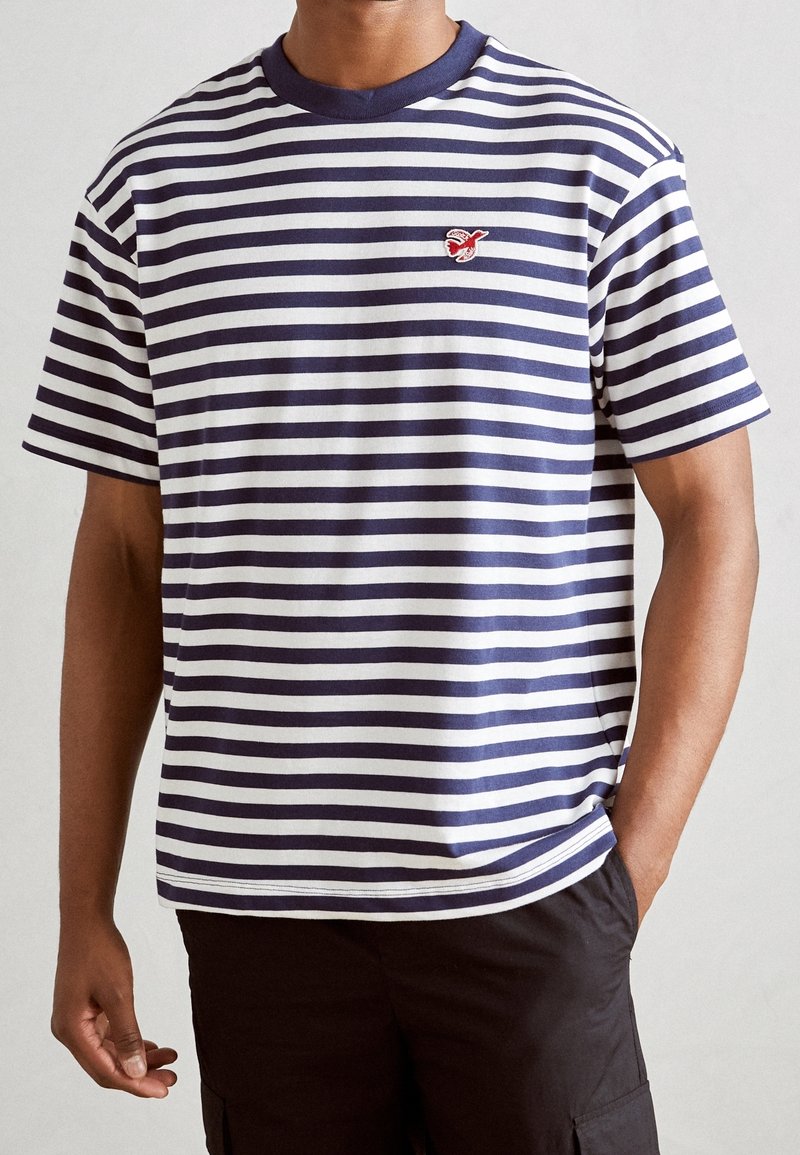 Stribet t-shirt med marineblå og hvide vandrette striber, ribbet marineblå krave og et lille rødt emblem på venstre bryst.