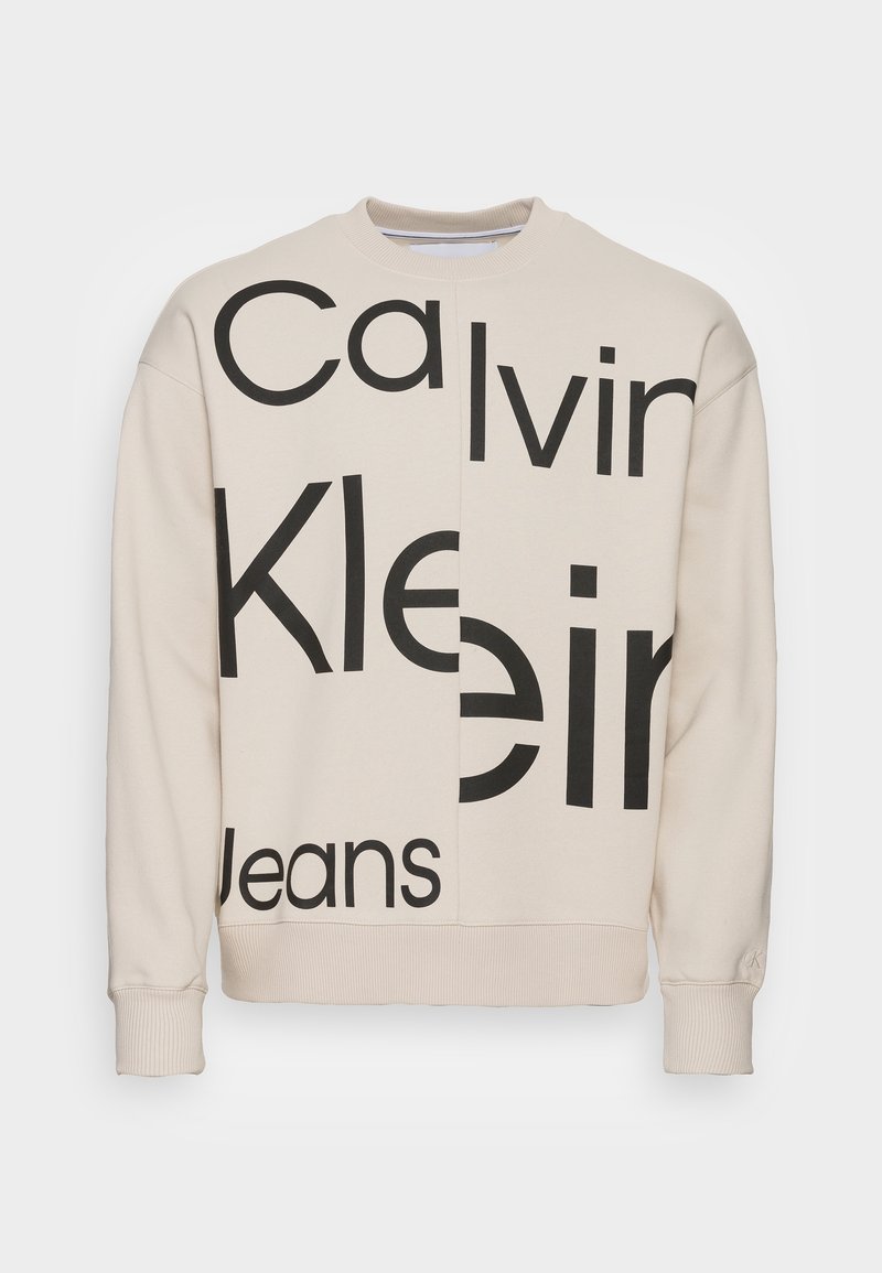 Sweatshirt beige à col rond avec un grand logo noir "Calvin Klein Jeans" imprimé verticalement au centre, poignets et ourlet côtelés.