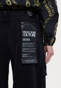 Črne denim hlače z vidnim logotipom v beli barvi. Stil vključuje žep z obližem in zapiranje na gumb.