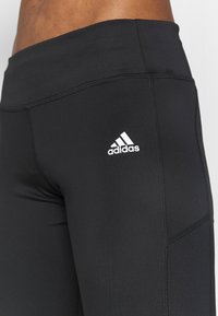 Černé atletické legíny z elastické látky. Mají hladkou texturu, vysoký pas a bílý logo Adidas na stehně.