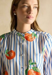 Bluse mit Knopfverschluss, die blau-weiße Streifen mit orangefarbigem Obst-Print zeigt, aus leichtem Stoff und mit Stehkragen. Mit strukturierten Details.
