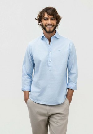 Joven sonriente con las manos en los bolsillos, vistiendo una camisa de manga larga azul claro y pantalones beige sobre un fondo liso.