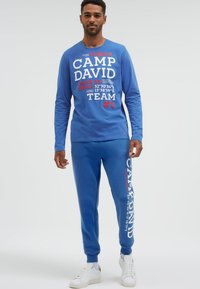 Mann steht, trägt blaues langärmeliges Hemd und Jogginghose mit weißem und rotem "Camp David"-Schriftzug, weiße Turnschuhe, vor einem schlichten hellen Hintergrund.