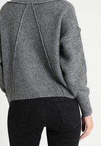 Szary sweter robiony na drutach o luźnym kroju, z prążkowanym dołem i szwem z tyłu. Zestawiony z czarnymi jeansami z wysokim stanem, posiadającymi tylne kieszenie.