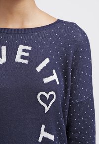 Marinblå stickad tröja med vit hjärta och text, rund halsringning, lös passform och subtil prickig mönster över hela tyget.