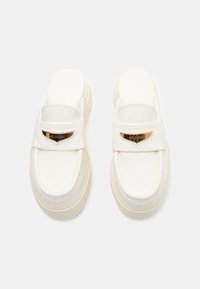 Et par hvite slip-on loafers med tykke såler og gullfargede "Air Jordan" plaketter på de øverste stroppene, vist mot en hvit bakgrunn.