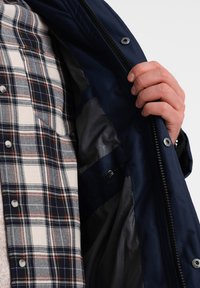 Námořnická modrá bunda se zapínáním na zip, která ukazuje kostkovanou košili uvnitř a hladké šedé podšívce. Obsahuje knoflíkové detaily a viditelné švy.