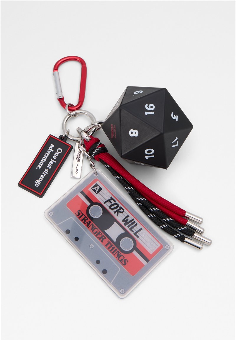 Porte-clés avec un mousqueton rouge, un dé noir à 20 faces, des cordons rouges et noirs, une étiquette cassette Stranger Things et une étiquette « Une dernière aventure étrange ».