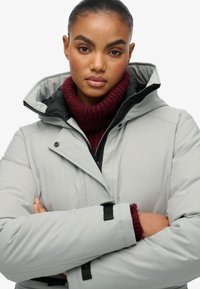 Młoda kobieta z założonymi rękami, ubrana w jasnoszary, zimowy płaszcz z kapturem oraz burgundowy, dzianinowy sweter z golfem.