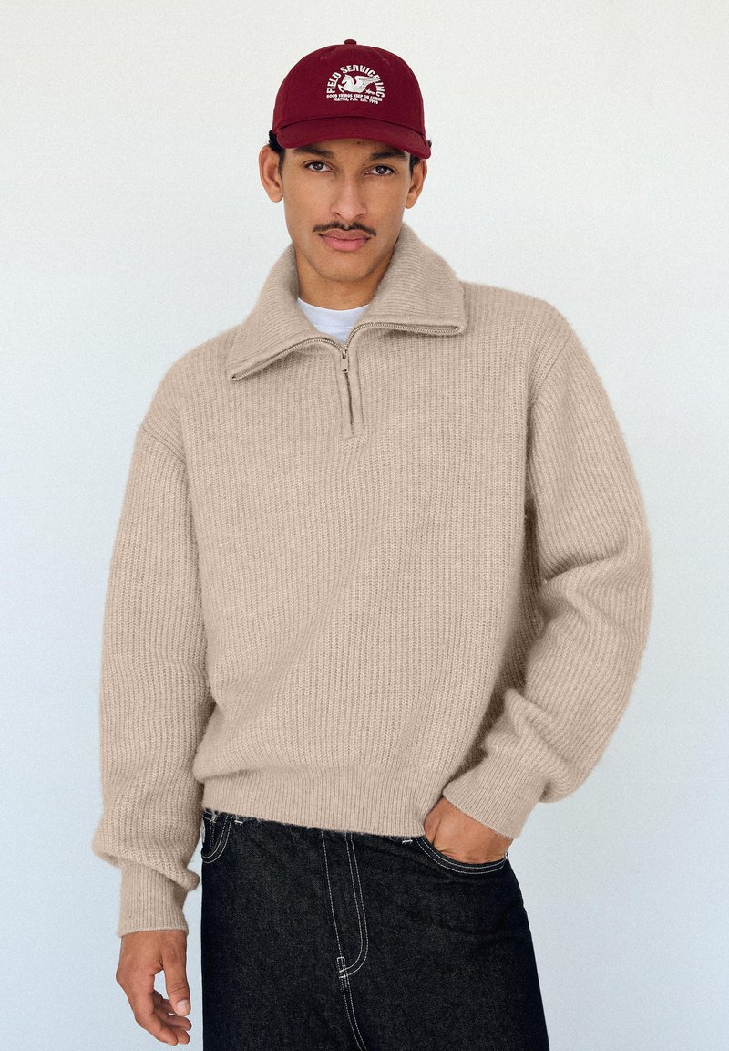 Maglione lavorato a maglia beige con collo con zip, maniche lunghe e texture a coste, abbinato a jeans in denim scuro e un berretto bordeaux.