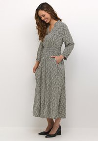 Duga haljina s uzorkom u crnoj i krem boji, s V-izrezom, dugim rukavima, elastičnom strukom i bočnim džepovima. Model nosi crne salonke.