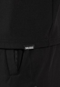 Nahaufnahme von schwarzem Stoff, der eine Saumnaht, einen Reißverschluss und ein kleines schwarzes Etikett mit weißer "SMILODOX"-Schrift zeigt.