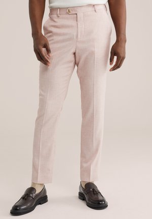 Muž v svetloružových na mieru ušitých nohaviciach, béžových ponožkách a tmavohnedých mokasínach s strapcami stojaci pred neutrálnym pozadím.