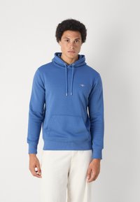 Jonge man die een blauwe pullover met capuchon, trekkoorden en een voorzak draagt, staand tegen een effen witte achtergrond.