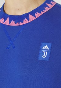 T-shirt de sport Adidas bleu avec écusson du logo Juventus sur la poitrine et motif triangulaire rose autour du col, porté par une personne.
