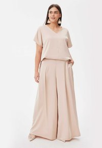 Beige twee-delige outfit met een losvallend T-shirt met korte mouwen en een V-hals, en wijde geplooide broeken. Soepele, zachte stof met zakken.