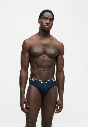 Homme musclé debout devant un fond uni, portant un slip BOSS bleu marine avec une ceinture blanche à logo, faisant face à l’avant avec une expression neutre.