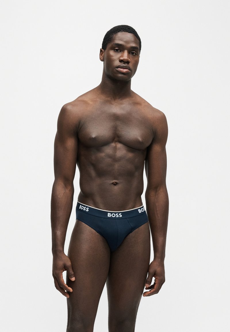 Homme musclé debout devant un fond uni, portant un slip BOSS bleu marine avec une ceinture blanche à logo, faisant face à l’avant avec une expression neutre.