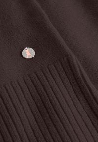 Bruine stof met een gestructureerd ribbelpatroon, voorzien van een cirkelvormige zilveren speld met een roze silhouetontwerp.