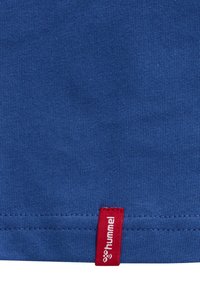 Tissu bleu avec une petite étiquette rouge cousue dans l'ourlet affichant le logo blanc "hummel" et l'icône d'abeille.