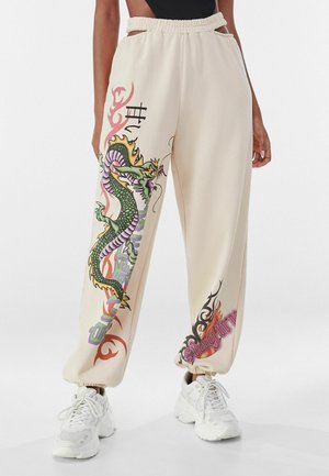 Luźne beżowe spodnie dresowe z kolorową zielono-fioletową grafiką smoka na lewej nogawce oraz napisem „Stay Tru” na prawym mankiecie, noszone z białymi sneakersami.