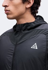 Jeune homme portant une veste noire zippée avec capuche et un logo triangulaire blanc ACG sur le côté gauche de la poitrine, visible du cou jusqu’au milieu de la poitrine.