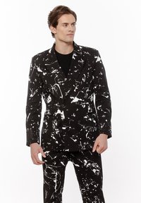 Młody mężczyzna ubrany w czarny, dwurzędowy garnitur z białym abstrakcyjnym wzorem plam farby, stojący na tle białym.