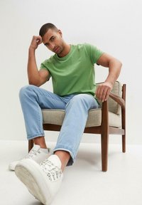 T-shirt verde a maniche corte, jeans dritti blu chiaro, e sneakers bianche con dettagli in metallo, seduto su una poltrona con telaio in legno.