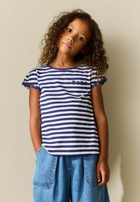 T-shirt à manches courtes rayé bleu marine et blanc avec une bordure florale délicate sur les manches et une petite poche poitrine. Porté avec un short en denim bleu clair.