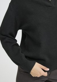 Juodas megztinis su V formos iškirpte. Minkšta tekstūra, gumbuotas apykaklė ir rankogaliai. Ranka poilsiauja ant pilkų kelnių kišenės. Švarus, minimalus dizainas.