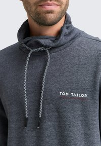 Grijze hoodie met koordjes, gemaakt van een gestructureerde stof, hoge kraag en een wit/rood "TOM TAILOR" logo op de voorkant.