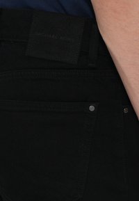 Black denim pants feature a leather-like label at the waistband and metal rivets on the front pockets. Smooth texture with a fitted design.