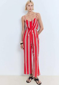 Une combinaison rayée rouge avec de fines bretelles, une taille cintrée et des jambes larges. Les rayures sont blanches et crème, créant un motif audacieux.